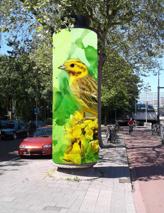 een aanplakzuil met handgeschilderde afbeelding van een geel vogeltje met groen en gele achtergrond